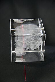The image shows a transparent acrylic cube. Inside the cube, there’s a white 3D-engraved model of a human brain. A laser beam creates a visible red line through the cube, illuminating part of the brain design. The cube is placed on a black surface.