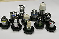 Photo of a series of microscope objectives of various types and designs. The objectives differ in magnification, size, and optical corrections, including achromatic, plan-achromatic, and apochromatic types. Some objectives are finite-corrected, others infinite-corrected. The photo illustrates the diverse components used for producing magnified images in light microscopy, highlighting the complexity and specialization of microscope optics