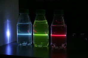 Image of a violet laser beam passing sequentially through three bottles containing different liquids. The liquid in the first bottle closest to the laser displays red fluorescence, the liquid in the middle bottle shows green fluorescence, and the liquid in the last bottle exhibits bright blue fluorescence. This setup demonstrates how distinct substances can emit different colors of fluorescent light when excited by the same laser, illustrating the principle of laser-induced fluorescence and energy conversion in various materials.