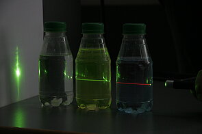 Image of a green laser beam passing sequentially through three bottles containing different liquids. The liquid in the first bottle closest to the laser displays red fluorescence, the liquid in the middle bottle shows green fluorescence, and the liquid in the last bottle exhibits no fluorescence. This setup demonstrates how distinct substances can emit different colors of fluorescent light when excited by the same laser, illustrating the principle of laser-induced fluorescence and energy conversion in various materials.