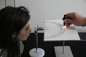 The image shows a scientific experiment involving two people. On the left, a woman is looking closely through a small cylindrical device mounted on a vertical stand, as if observing something through a lens. In front of her, horizontally placed, is a sheet of paper or plastic printed with a large semicircular protractor marked in degrees from 0 to 360 at regular intervals. To the right, another person’s hand holds a small flashlight or laser pointer, directing a narrow beam of light onto the protractor near the 220-degree mark. 