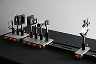 The image shows five optical components arranged in a row on a black surface. Each component is mounted on an adjustable gray stand with an orange knob. From left to right, the components are: a large circular lens/mirror assembly, a square dichroic mirror/filter, a smaller circular convex lens, another square lens/filter, and a small black light source module. The stands are attached to a long black base, and the setup is positioned against a light gray wall. 