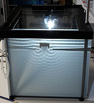 Image of a ripple tank, a shallow transparent tray filled with water used to study wave behavior. The tank shows waves generated on the water surface, illustrating phenomena such as reflection, refraction, diffraction, and interference. Light shining through or above the water enhances the visibility of wave patterns.