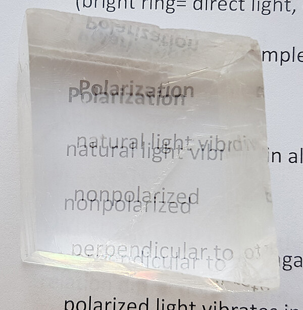 Image of a transparent calcite crystal resting on a printed text, causing the letters beneath to appear doubled. This optical effect, known as birefringence or double refraction, occurs when the calcite splits incoming light into two rays, making everything viewed through the crystal look as if it is duplicated. The phenomenon demonstrates the unique optical property of calcite, often used for educational purposes to illustrate light refraction and polarization.
