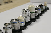 Photo of a series of microscope objectives of various types and designs. The objectives differ in magnification, size, and optical corrections, including achromatic, plan-achromatic, and apochromatic types. Some objectives are finite-corrected, others infinite-corrected. The photo illustrates the diverse components used for producing magnified images in light microscopy, highlighting the complexity and specialization of microscope optics