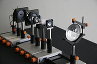 The image displays a setup of multiple optical components arranged in a straight line on a black table. Each component is mounted on a sturdy metal base and held upright by vertical rods. The devices include several black frames with circular openings (likely optical apertures or diaphragms), one large round glass lens, and smaller lenses or glass elements positioned between the frames. Several orange knobs are used for tightening or adjusting the mounts. The overall setup is precise and organized, resembling equipment used for physics experiments with light, such as a demonstration of lenses or an optics bench. 