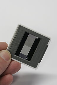 The image shows a close-up of a rectangular object held between fingers against a plain light background. The object is a dia slide with a gray frame and a black center. In the center of the slide, there are several horizontal bands: the topmost band has fine vertical lines or stripes,  and below it are several solid gray sections ranging from light to dark.  The letter "C" is marked in the upper right corner, and "F" is in the lower left. 