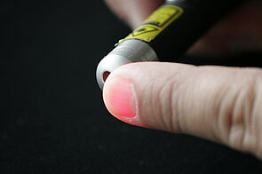 The image shows a close-up of a person's fingertip being touched by a small, cylindrical device that emits a red light. The device is small and cylindrical, with a metallic finish. It has a yellow label with black text, though the text is not legible. A red light is emitted from the tip of the device, illuminating the fingertip.