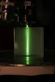 The image shows a close-up view of a scientific setup, possibly a microscope, with a rectangular, transparent block resting on a reflective surface. A bright green light is emanating from the bottom of the block, shining upwards through it. The light creates a distinct, vertical green line that extends upwards in a narrow angle.
